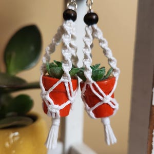 Op de afbeelding: Een paar macramé plantenhanger oorbellen met kleine oranje potten en groene planten. De oorbellen hebben houten kralen en zilveren haken.