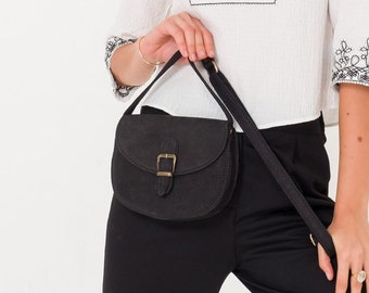 Puede incluir: Una mujer lleva una blusa blanca bordada con ribetes negros y pantalones negros. Lleva un bolso de cuero negro con una hebilla dorada.