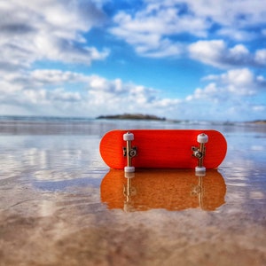 Puede incluir: Un monopatín en miniatura de color naranja brillante, con ruedas blancas, descansa sobre arena mojada, reflejando el cielo azul y nublado. El contraste del monopatín destaca sobre el fondo de la playa.