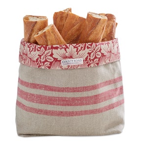 May include: A beige linen bread basket with red and white floral trim and red stripes. The basket is filled with several baguettes. The basket has a label that reads "COUNTY ROAD - AMERICAN MADE".