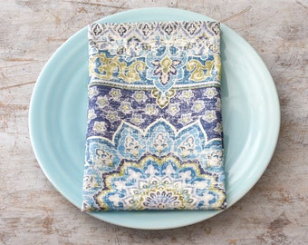May include: A blue and white patterned fabric napkin folded and placed on a light blue plate. The plate is on a light wood surface.