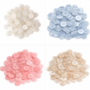 May include: Four piles of decorative buttons in various colours. The top left pile is white, the top right is light blue, the bottom left is pink, and the bottom right is beige. Each button has a two-hole design.