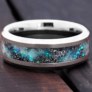 Puede incluir: Un anillo plateado con una banda de incrustaciones brillantes azules y verdes. El anillo tiene un acabado metálico pulido y un interior oscuro y texturizado. La incrustación tiene un diseño cósmico moteado.