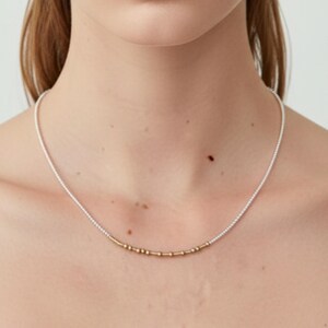 Op de afbeelding: Een delicate ketting met een lange streng kleine witte kralen, geaccentueerd met een centraal gedeelte van kleine, cilindrische goudkleurige kralen. De ketting rust tegen de hals en heeft een minimalistisch ontwerp met een gladde textuur.