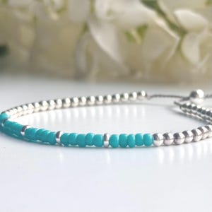 Op de afbeelding: Een delicate armband met een herhalend patroon van kleine, ronde, metallic zilverkleurige kralen afgewisseld met levendige turquoise zaadkralen. Het ontwerp bevat een verstelbaar schuifsluitingsmechanisme met zilverkleurige kralen.