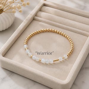 Puede incluir: Una pulsera circular de cuentas que alterna pequeñas esferas de tono dorado metálico y cuentas blancas translúcidas. La pulsera incluye una secuencia de cuentas dispuestas en un patrón con la palabra "Warrior" impresa en una fuente oscura.