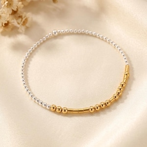 May include: A delicate bracelet featuring a circular arrangement of small, polished silver-toned beads along the majority of the band, transitioning into a segment of gold-toned beads and cylindrical spacers arranged in a linear pattern.
