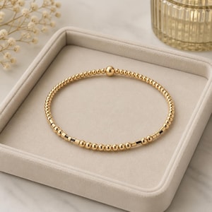 May include: A gold beaded bracelet featuring a series of small, round, polished gold-tone beads arranged in a circular band. The design includes a repeating pattern of gold beads interspersed with small, dark, rectangular spacers along the circumference.