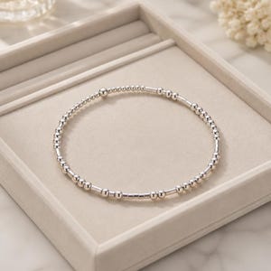 May include: A delicate silver-toned bracelet composed of a repeating pattern of small round beads and short cylindrical tube beads. The metallic accessory features a continuous circular design with a polished finish, creating a simple and uniform look.
