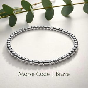 Könnte beinhalten: Ein silbernes Perlenarmband mit den Worten "Morse Code | Brave" in einer Serifenschrift. Das Armband besteht aus kleinen, runden Perlen und länglichen Perlen. Grüne Blätter sind im Hintergrund.