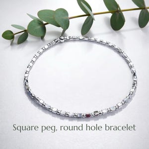 Puede incluir: Un brazalete plateado con cuentas cuadradas y redondas. Se muestra sobre un fondo blanco, con una ramita de hojas verdes. El texto "Square peg, round hole bracelet" es visible.