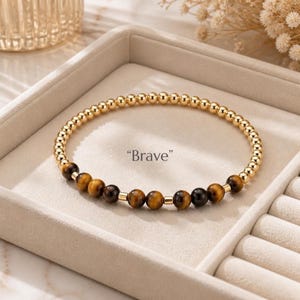 Puede incluir: Una pulsera de cuentas que presenta una larga hilera de pequeñas cuentas metálicas en tono dorado y una sección de cuentas de piedra preciosa ojo de tigre en marrón y negro. La palabra "Brave" está impresa en el centro. Diseño circular.