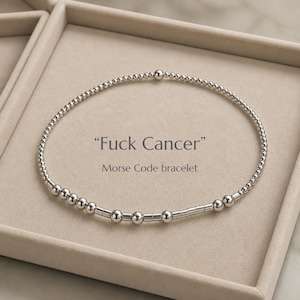 Pode incluir: Uma pulseira de contas prateadas com um padrão em código Morse. O texto "Fuck Cancer" e "Morse Code bracelet" está impresso no centro. A pulseira é composta por pequenas contas redondas e espaçadores cilíndricos curtos dispostos em círculo.