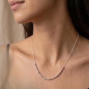 Op de afbeelding: Een zilverkleurige ketting met een fijne schakelketting en een centraal staafje versierd met een reeks kleine, verhoogde bolvormige kralen en gladde cilindrische segmenten in een lineair patroon over het sleutelbeen.