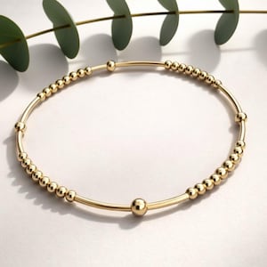 May include: A gold bracelet featuring small, round beads threaded onto a curved, slender band. The bracelet is displayed against a white backdrop, with a sprig of green leaves in the upper left. A delicate piece of jewellery.