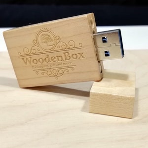 Può includere: Un'unità USB in legno con una finitura in legno chiaro. L'unità è incisa con il testo "WoodenBox Packaging, gift and more..." e un design stilizzato di un albero. L'unità è aperta e il connettore USB è visibile.