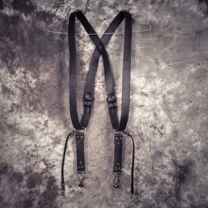 May include: Black leather camera strap with adjustable straps and metal hardware. The strap is hanging on a hanger.