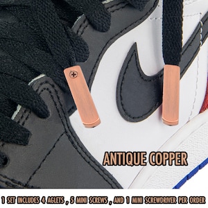 May include: Close-up of a sneaker with black laces and antique copper aglets. The shoe has a white and black leather design with a black Nike swoosh. The text "ANTIQUE COPPER" is visible. The set includes 4 aglets, 5 mini screws, and 1 mini screwdriver.