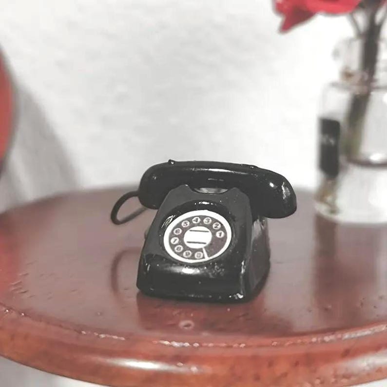 Puede incluir: Un peque&ntilde;o tel&eacute;fono rotatorio negro con un dial blanco y un auricular curvo. El tel&eacute;fono est&aacute; sobre una superficie de madera oscura. Un jarr&oacute;n con flores rojas est&aacute; en el fondo.