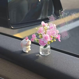May include: A small white rabbit figurine with a pink butterfly on its head sits next to a clear glass bottle containing a bouquet of pink artificial flowers. The bottle features a small white label with the text GOOD LUCK printed in black.