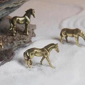 Puede incluir: Cuatro peque&ntilde;as figuras de caballos dorados en varias poses. Una est&aacute; sobre una roca, mientras que otras est&aacute;n sobre una superficie blanca y texturizada. Los caballos son detallados, con crines y colas visibles, lo que sugiere una colecci&oacute;n de esculturas en miniatura.