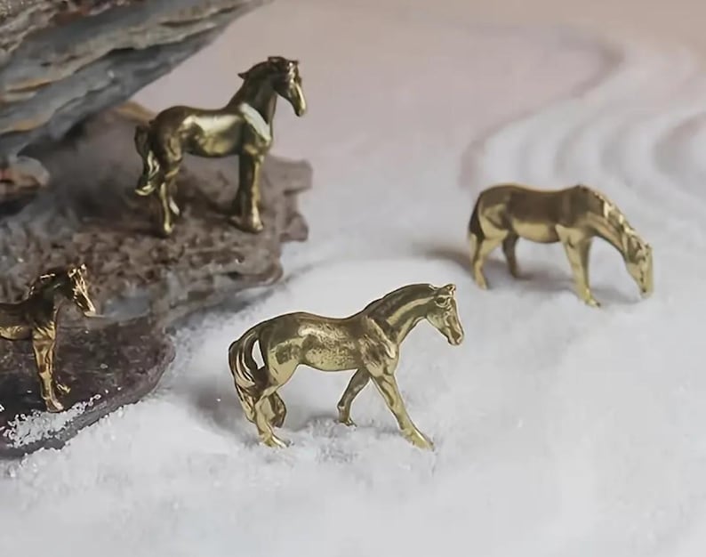 Puede incluir: Cuatro peque&ntilde;as figuras de caballos dorados en varias poses. Una est&aacute; sobre una roca, mientras que otras est&aacute;n sobre una superficie blanca y texturizada. Los caballos son detallados, con crines y colas visibles, lo que sugiere una colecci&oacute;n de esculturas en miniatura.