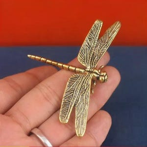 May include: A metallic gold-toned dragonfly figurine featuring intricate, etched wing patterns and a segmented body. The insect sculpture displays a polished finish with dark recessed detailing in the wings, showcasing a realistic form with four extended wings.