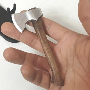 May include: A miniature axe with a polished steel blade and a dark wooden handle. The small axe is held in a hand, showcasing the size and craftsmanship of the tool. The axe is a miniature version of a larger tool.