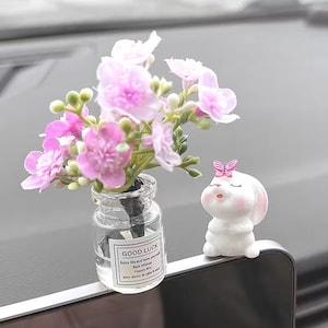 May include: A small glass vase with pink flowers and a white bunny figurine with a pink butterfly on its head. The vase has a label with the words "Good Luck" and other text. The bunny is sitting down.