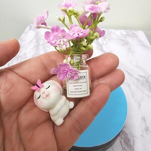 May include: A miniature decorative arrangement featuring a small glass bottle with pink flowers and the word "LUCK" on a label. A small white bunny figurine with a pink butterfly sits nearby. The arrangement is held in a hand.