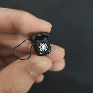 Puede incluir: Un peque&ntilde;o tel&eacute;fono rotatorio negro con un dial blanco y un cable negro. El tel&eacute;fono en miniatura de estilo vintage se sostiene en una mano sobre un fondo oscuro. El tel&eacute;fono es una r&eacute;plica de un dise&ntilde;o cl&aacute;sico.