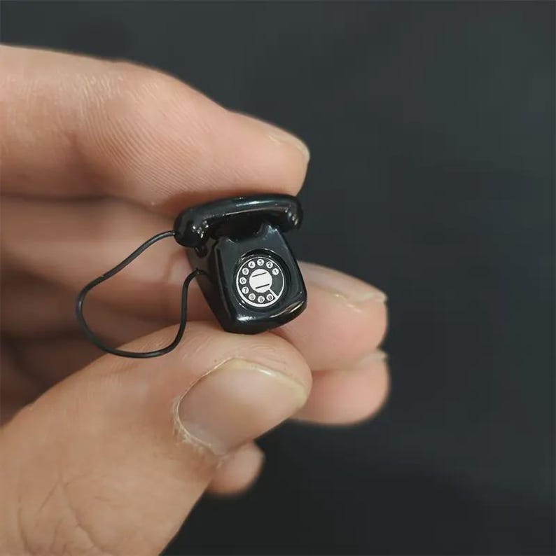 Puede incluir: Un peque&ntilde;o tel&eacute;fono rotatorio negro con un dial blanco y un cable negro. El tel&eacute;fono en miniatura de estilo vintage se sostiene en una mano sobre un fondo oscuro. El tel&eacute;fono es una r&eacute;plica de un dise&ntilde;o cl&aacute;sico.