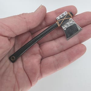 May include: A miniature axe with a black handle and a silver-colored head. The head has a textured pattern and is bound to the handle with twine. The axe is held in a person's hand against a white background.