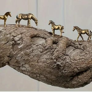 Puede incluir: Cuatro peque&ntilde;as figuras de caballos dorados est&aacute;n dispuestas sobre una rama de madera marr&oacute;n texturizada. Los caballos est&aacute;n en varias poses, incluyendo de pie y pastando. La rama tiene una apariencia rugosa, similar a la corteza.