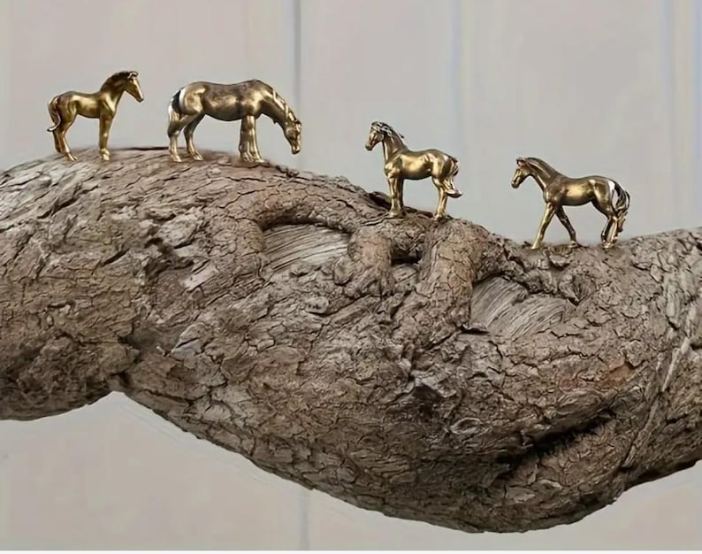 Puede incluir: Cuatro peque&ntilde;as figuras de caballos dorados est&aacute;n dispuestas sobre una rama de madera marr&oacute;n texturizada. Los caballos est&aacute;n en varias poses, incluyendo de pie y pastando. La rama tiene una apariencia rugosa, similar a la corteza.