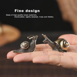 May include: Two brass snail figurines. The snails are intricately detailed with textured bodies and spiraled shells. The text "Fine design" and other descriptive text is visible in the background.