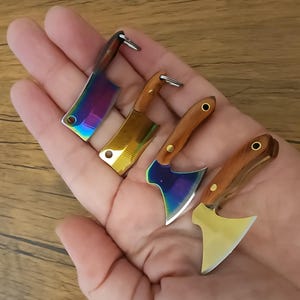 May include: Four miniature metal cleavers and axes with wooden handles are displayed. The blades have iridescent, gold, and silver finishes. The cleavers have a rectangular shape, while the axes have a curved blade.
