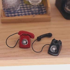 Puede incluir: Miniaturas de tel&eacute;fonos rotatorios rojos y negros con cables negros. El tel&eacute;fono rojo tiene un auricular y est&aacute; al lado de un tel&eacute;fono negro con un auricular. Los tel&eacute;fonos est&aacute;n sobre una superficie de madera.