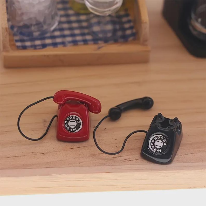 Puede incluir: Miniaturas de tel&eacute;fonos rotatorios rojos y negros con cables negros. El tel&eacute;fono rojo tiene un auricular y est&aacute; al lado de un tel&eacute;fono negro con un auricular. Los tel&eacute;fonos est&aacute;n sobre una superficie de madera.