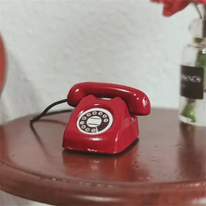 Puede incluir: Un tel&eacute;fono rojo en miniatura con un cable negro se encuentra sobre una mesa marr&oacute;n oscuro. El tel&eacute;fono tiene un dial circular con n&uacute;meros blancos y un auricular rojo a juego. El fondo incluye una pared blanca y un jarr&oacute;n de cristal.