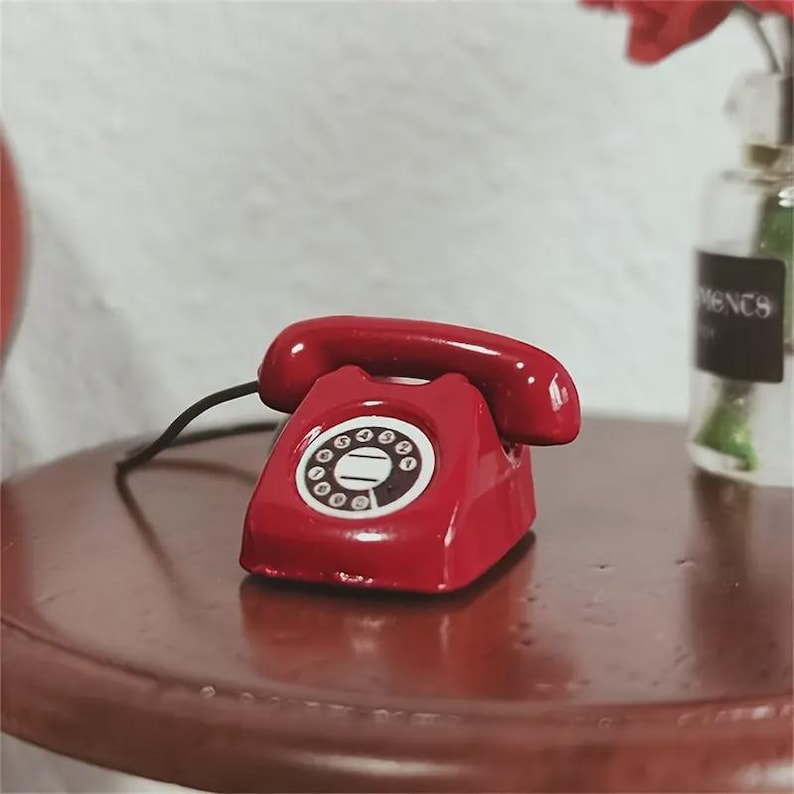 Puede incluir: Un tel&eacute;fono rojo en miniatura con un cable negro se encuentra sobre una mesa marr&oacute;n oscuro. El tel&eacute;fono tiene un dial circular con n&uacute;meros blancos y un auricular rojo a juego. El fondo incluye una pared blanca y un jarr&oacute;n de cristal.