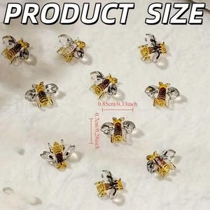 May include: Ten small bee-shaped charms with amber-colored bodies and clear wings. The text PRODUCT SIZE is at the top. A measurement indicator shows the bees are 0.85 cm wide and 0.7 cm tall, which is approximately 0.33 inches by 0.28 inches.