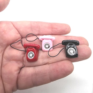 Puede incluir: Tres tel&eacute;fonos rotatorios en miniatura en rojo, rosa y negro. Cada tel&eacute;fono tiene un dial y un auricular conectados por un cable negro. Los tel&eacute;fonos son lo suficientemente peque&ntilde;os como para caber en la palma de una mano.