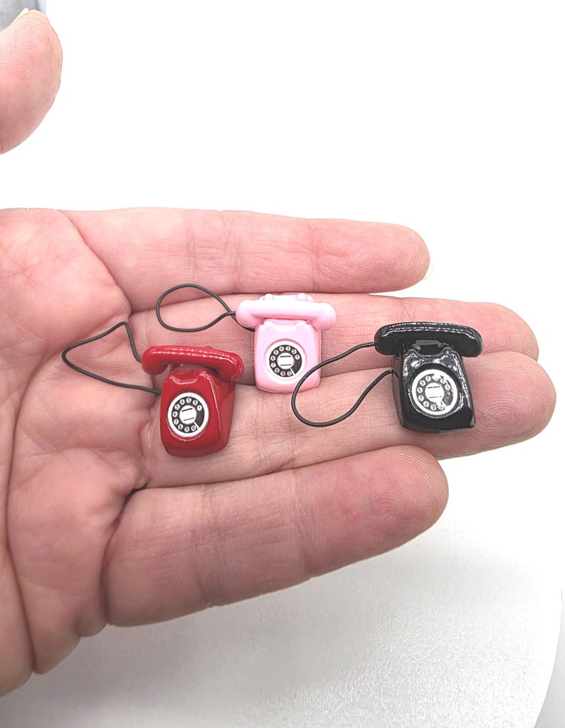 Puede incluir: Tres tel&eacute;fonos rotatorios en miniatura en rojo, rosa y negro. Cada tel&eacute;fono tiene un dial y un auricular conectados por un cable negro. Los tel&eacute;fonos son lo suficientemente peque&ntilde;os como para caber en la palma de una mano.