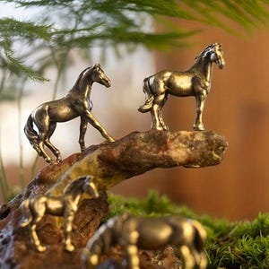 Puede incluir: Varias figuras de caballos de color bronce se colocan sobre una base de madera marr&oacute;n texturizada, algunas en movimiento y otras de pie. Las figuras se colocan sobre un fondo de vegetaci&oacute;n y un fondo borroso de tonos c&aacute;lidos.