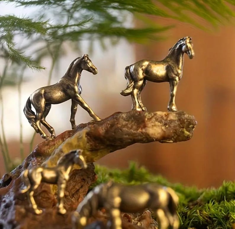 Puede incluir: Varias figuras de caballos de color bronce se colocan sobre una base de madera marr&oacute;n texturizada, algunas en movimiento y otras de pie. Las figuras se colocan sobre un fondo de vegetaci&oacute;n y un fondo borroso de tonos c&aacute;lidos.