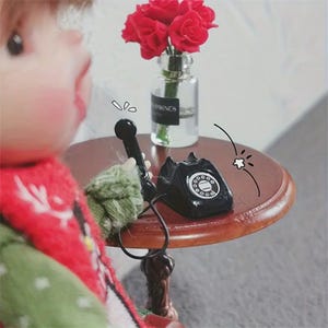 Puede incluir: Una escena en miniatura con una peque&ntilde;a mesa redonda marr&oacute;n. Sobre la mesa, un tel&eacute;fono rotatorio negro y un auricular negro. Un peque&ntilde;o jarr&oacute;n de cristal contiene rosas rojas. Accesorio de casa de mu&ntilde;ecas.