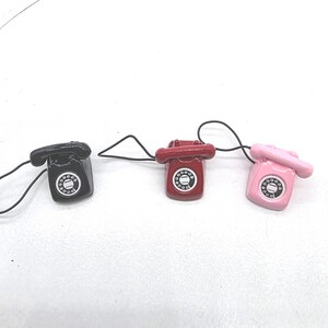Puede incluir: Tres tel&eacute;fonos en miniatura de estilo retro en negro, rojo y rosa. Cada tel&eacute;fono tiene un dial giratorio y un cable negro. Los tel&eacute;fonos son peque&ntilde;os, probablemente amuletos o art&iacute;culos decorativos.