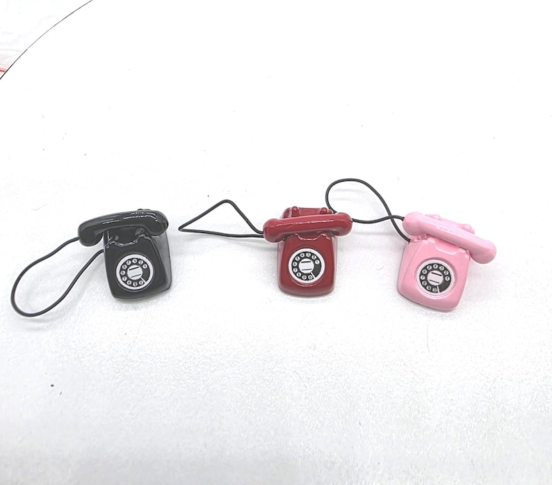 Puede incluir: Tres tel&eacute;fonos en miniatura de estilo retro en negro, rojo y rosa. Cada tel&eacute;fono tiene un dial giratorio y un cable negro. Los tel&eacute;fonos son peque&ntilde;os, probablemente amuletos o art&iacute;culos decorativos.