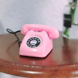 Puede incluir: Un tel&eacute;fono rotatorio rosa en miniatura con un dial negro y un auricular a juego. El tel&eacute;fono est&aacute; sobre una superficie marr&oacute;n, con un cable negro. El dise&ntilde;o recuerda a los tel&eacute;fonos antiguos.